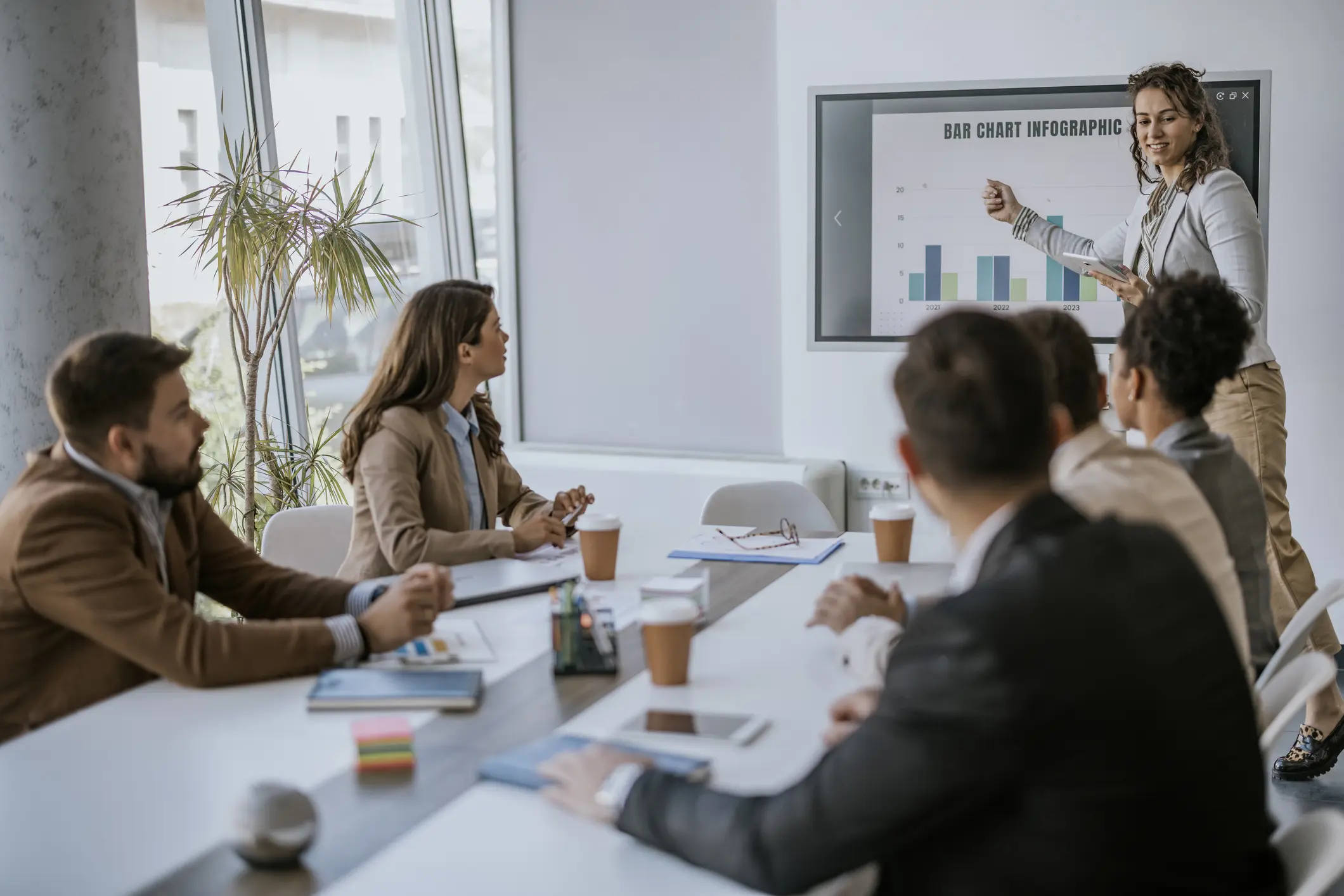 A businesswoman stands in front of a modern office conference table, presenting a bar chart infographic to her team. The presentation is focused on the bar chart, which is displayed on a screen on the wall. The team members are attentive and engaged in the presentation.