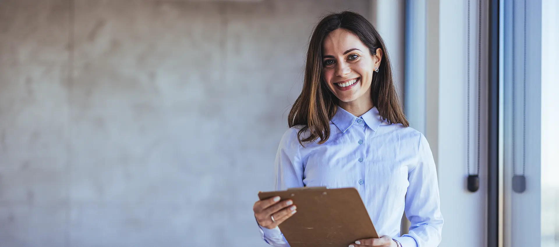 Smiling woman holding clipboard near window.